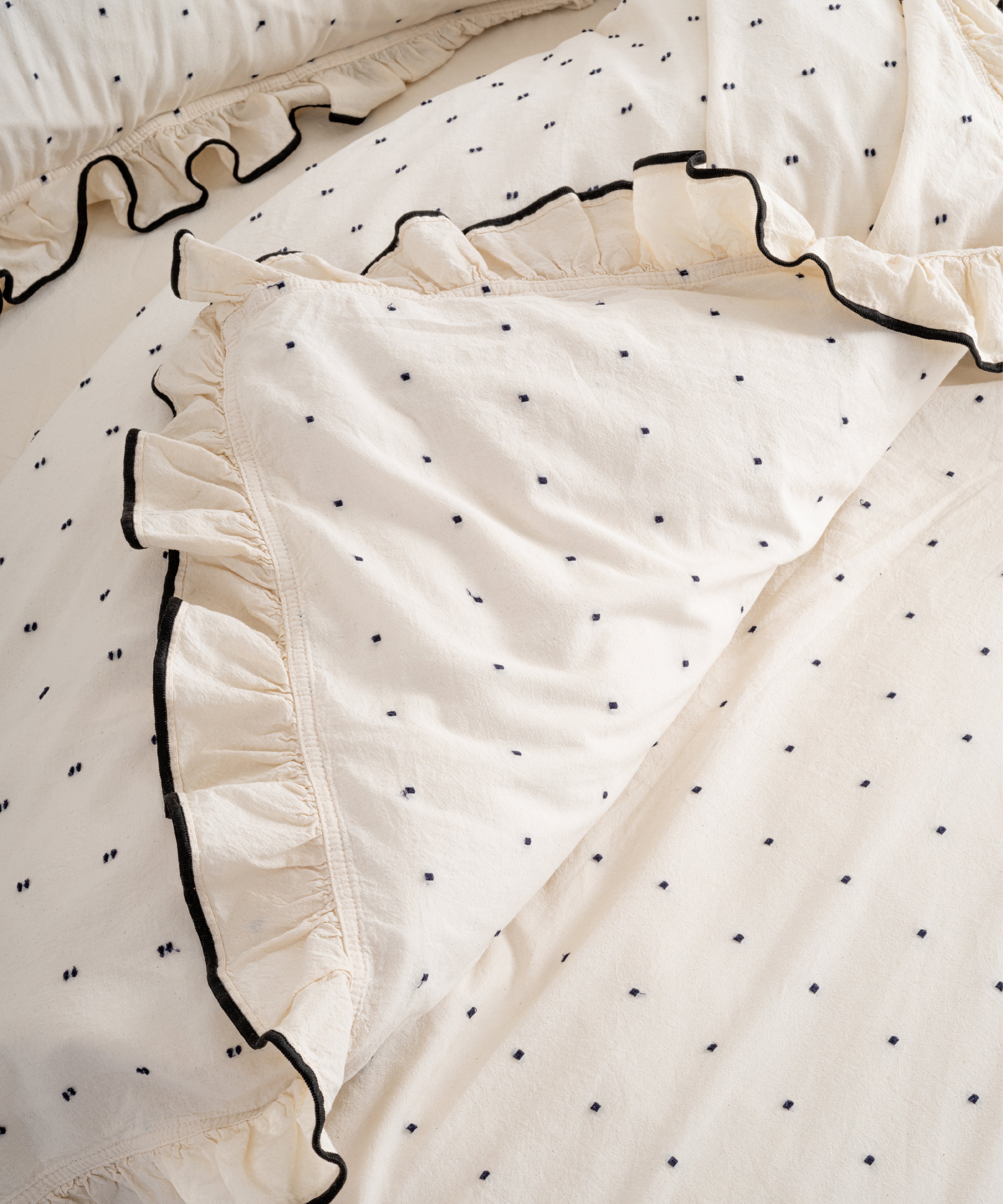 Top-down view of Limasso London Point Exclusive garment-washed cotton duvet cover set – cream with black micro-dots and black embroidered ruffle trim, open fold detail