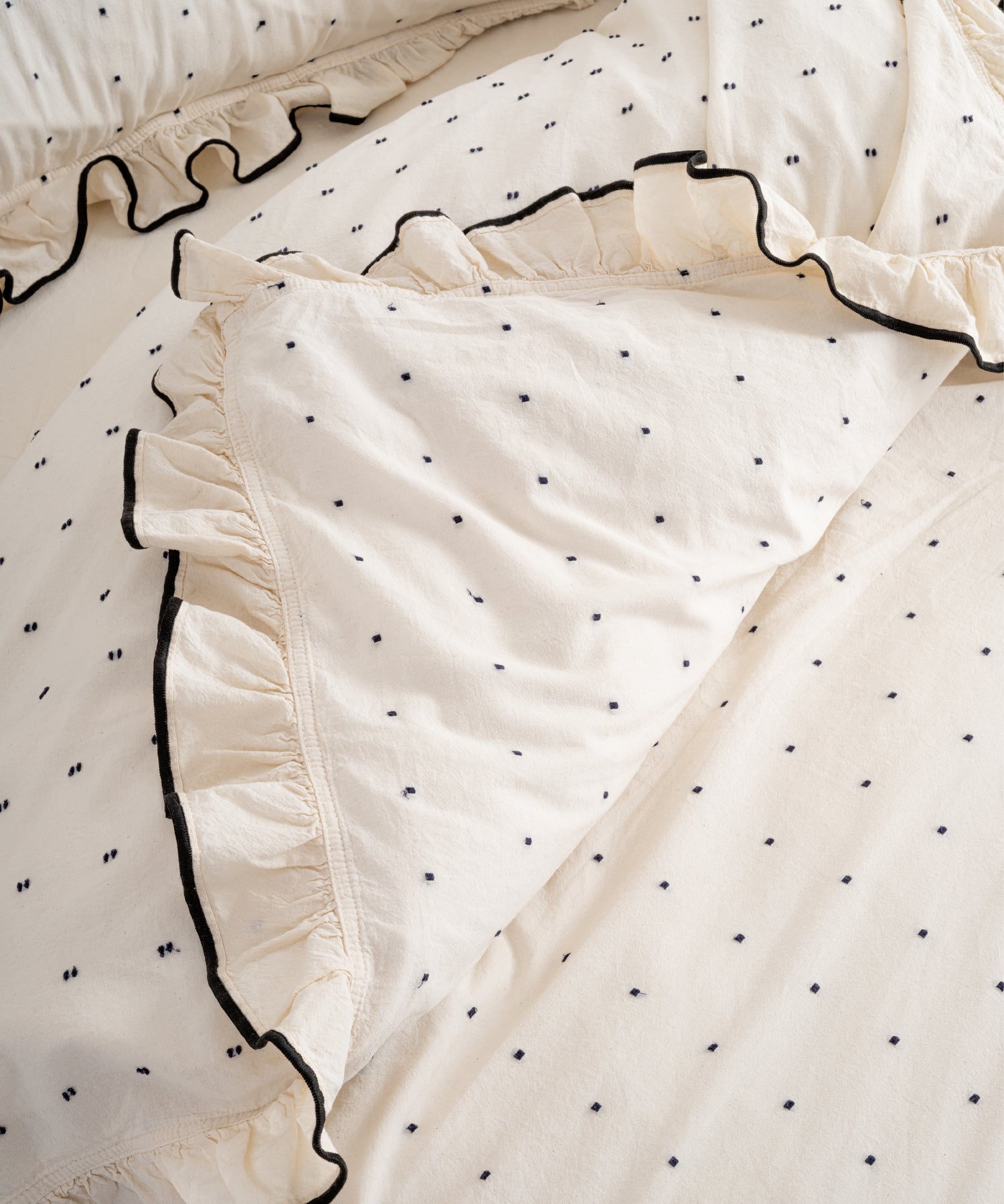 Top-down view of Limasso London Point Exclusive garment-washed cotton duvet cover set – cream with black micro-dots and black embroidered ruffle trim, open fold detail
